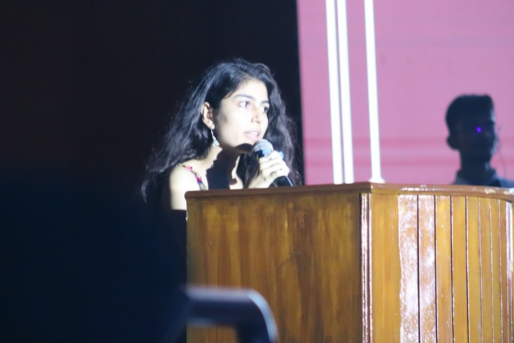 Diya speaking at a podium on a dark stage